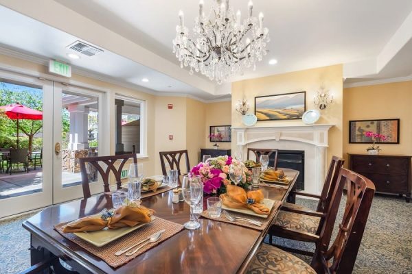 A dining room with a large table and chairs in front of a fireplace. There are lots of windows and a fireplace.