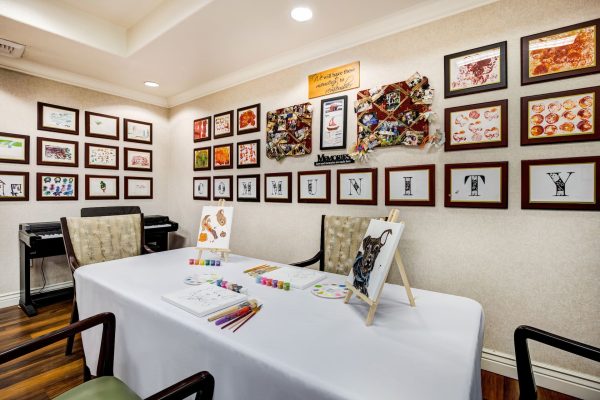 A painting table and chairs with art supplies on it and residents’ art hanging on the wall in the background.