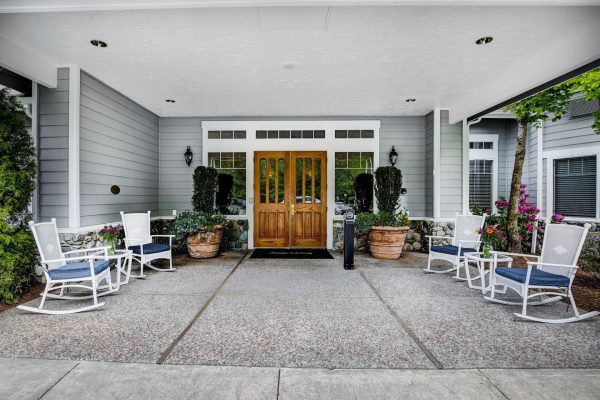 A covered outdoor area with four white rocking chairs and two small tables.