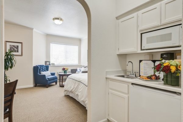 A small kitchen with flowers on the counter, with a view of the bedroom in the background