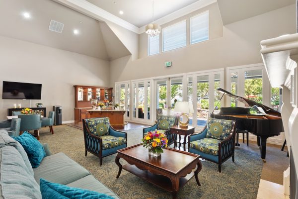 A large seating area with many windows, a couch, tables and armchairs, and a piano.