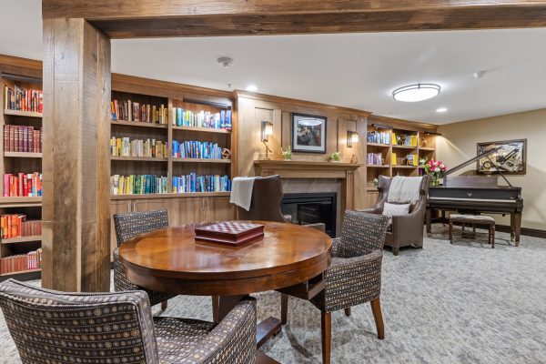 A library with a built-in bookshelf, tables, chairs, a piano, and a fireplace