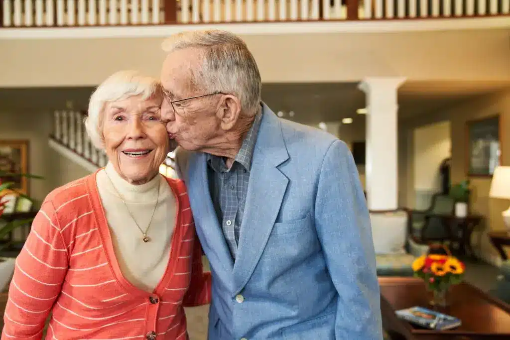 Elderly couple in living room, man kisses woman's cheek.